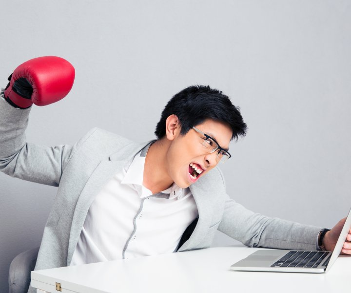 Angry young businessman sitting at the table and reading to hit the laptop over gray background