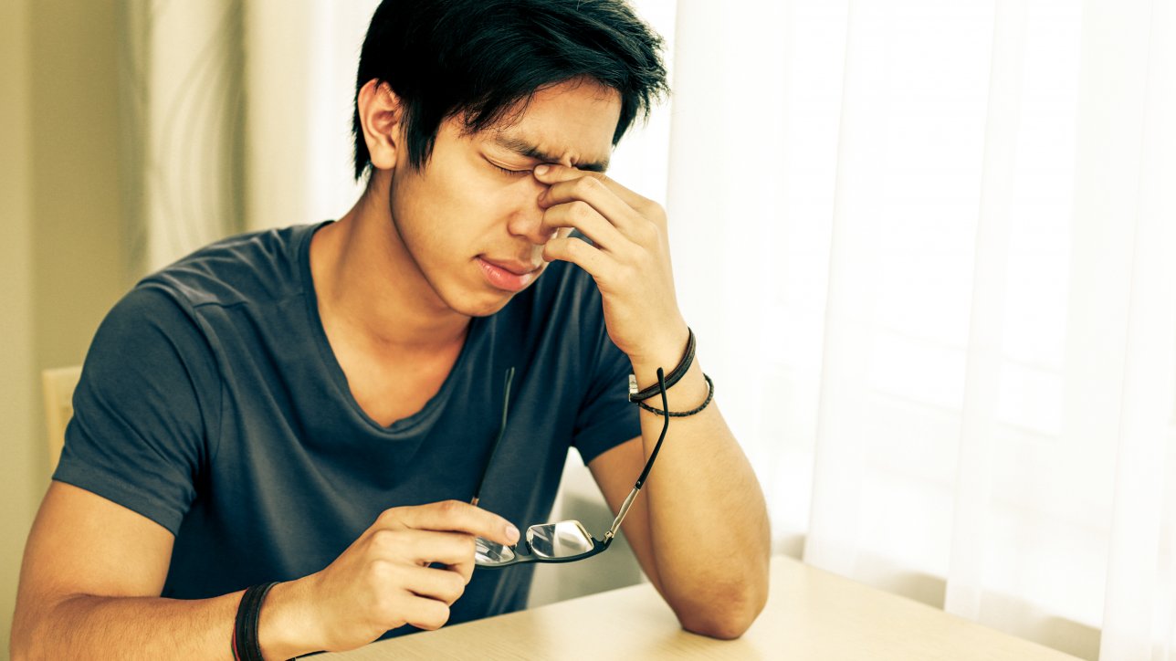 Tired asian man with eye pain holding glasses in hand
