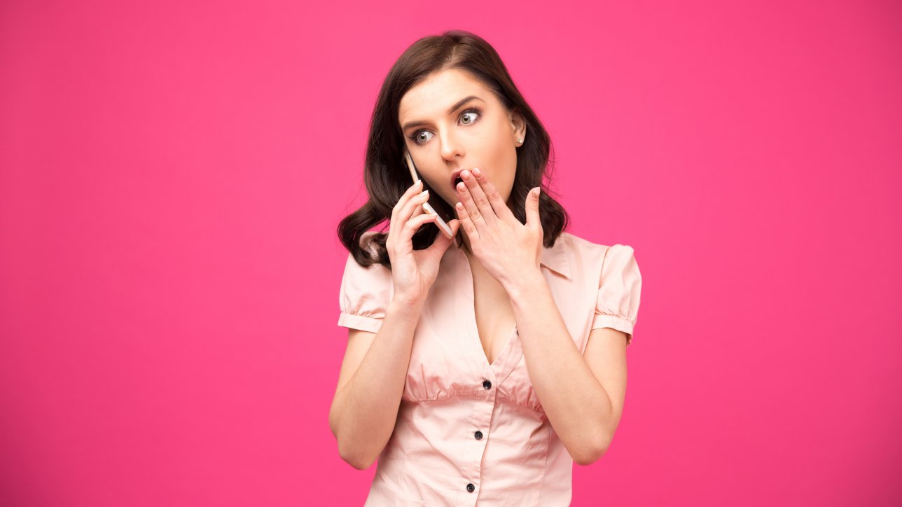 Surprised young woman talking on the phone over pink background