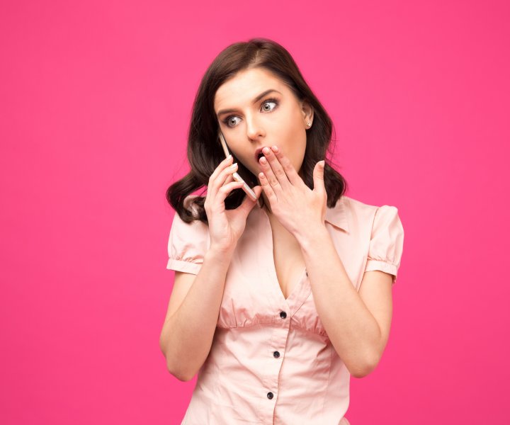 Surprised young woman talking on the phone over pink background